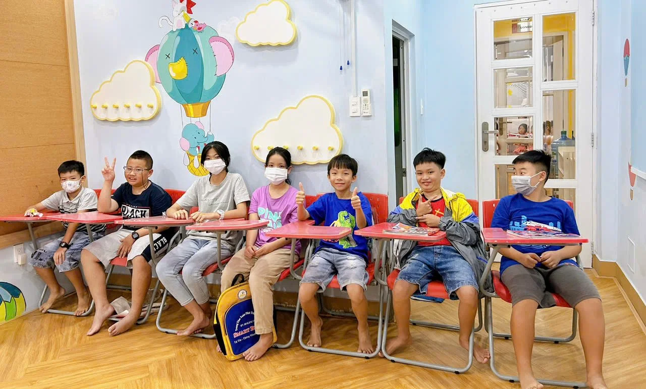 Seven children sitting at red desks in a colorful room with cloud and animal wall decorations, all wearing masks.