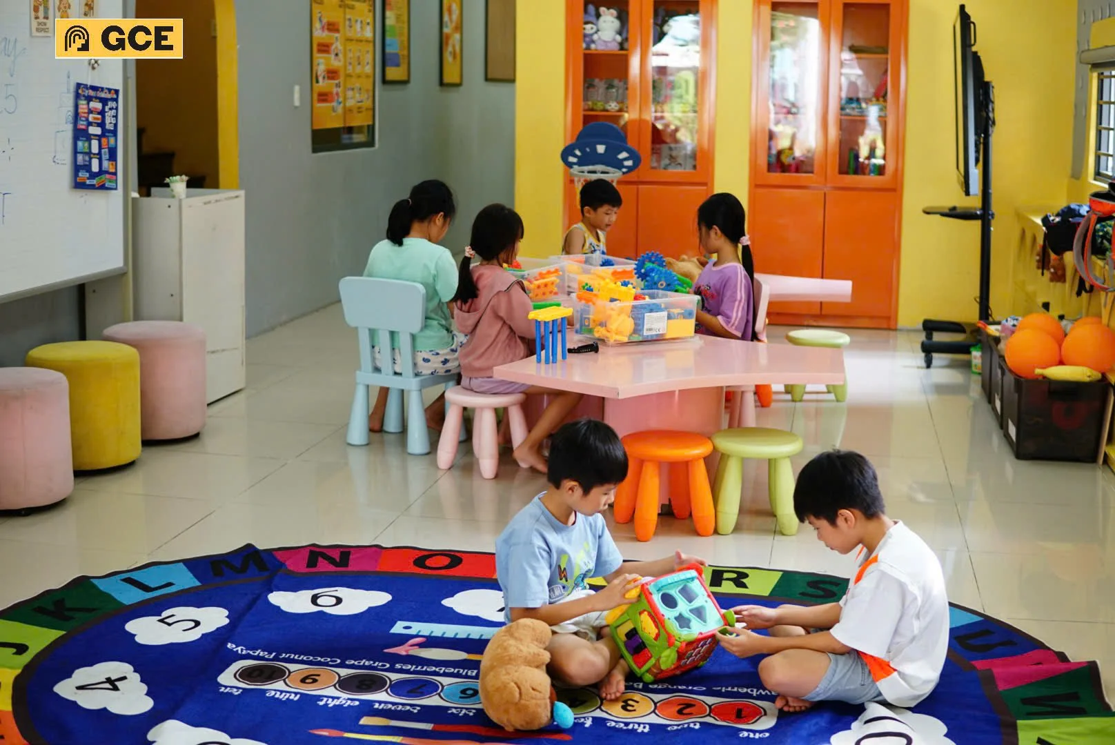 Children playing on a colorful alphabet and numbers rug in a classroom or daycare setting with tables, chairs, toys, and storage cabinets.