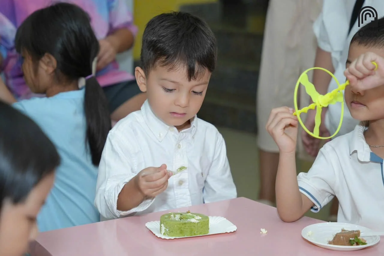 Children sitting at a table, with one boy examining a piece of cake and another boy holding a yellow toy helicopter.
