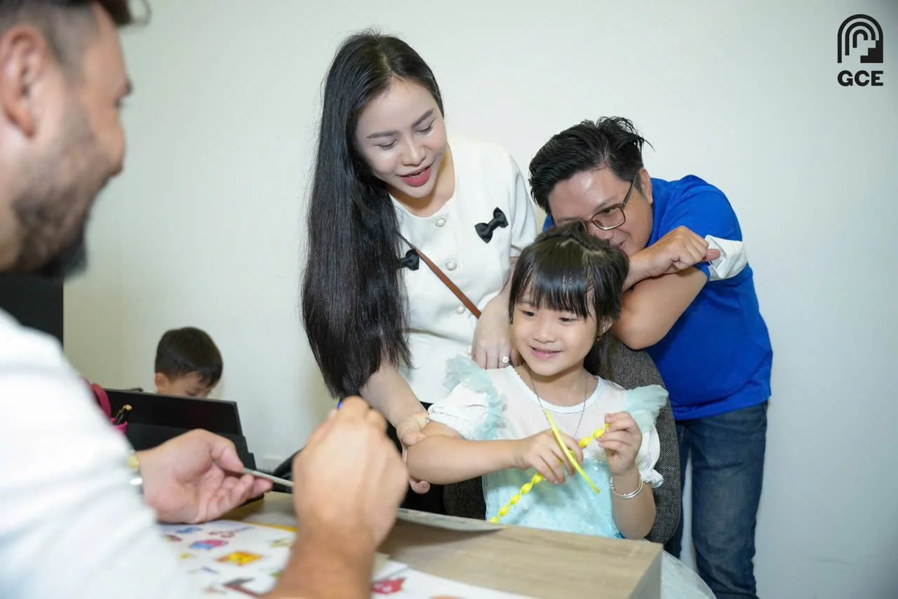 A young girl with dark hair and a light blue dress is smiling while holding yellow, twisted paper craft. She is sitting at a table, and a woman and a man are playfully pulling her arms. The woman has long black hair and is wearing a white blouse with black bow details, and the man has short dark hair, glasses, and is wearing a blue shirt. In the background, an older man with gray hair and a beard, wearing a white shirt, is working on something at the table. There is another child in the background sitting at the table as well.