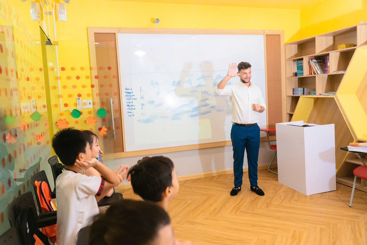 A classroom with a teacher standing in front of a whiteboard, waving, and students sitting on chairs facing him, with a yellow wall and bookshelves in the background.
