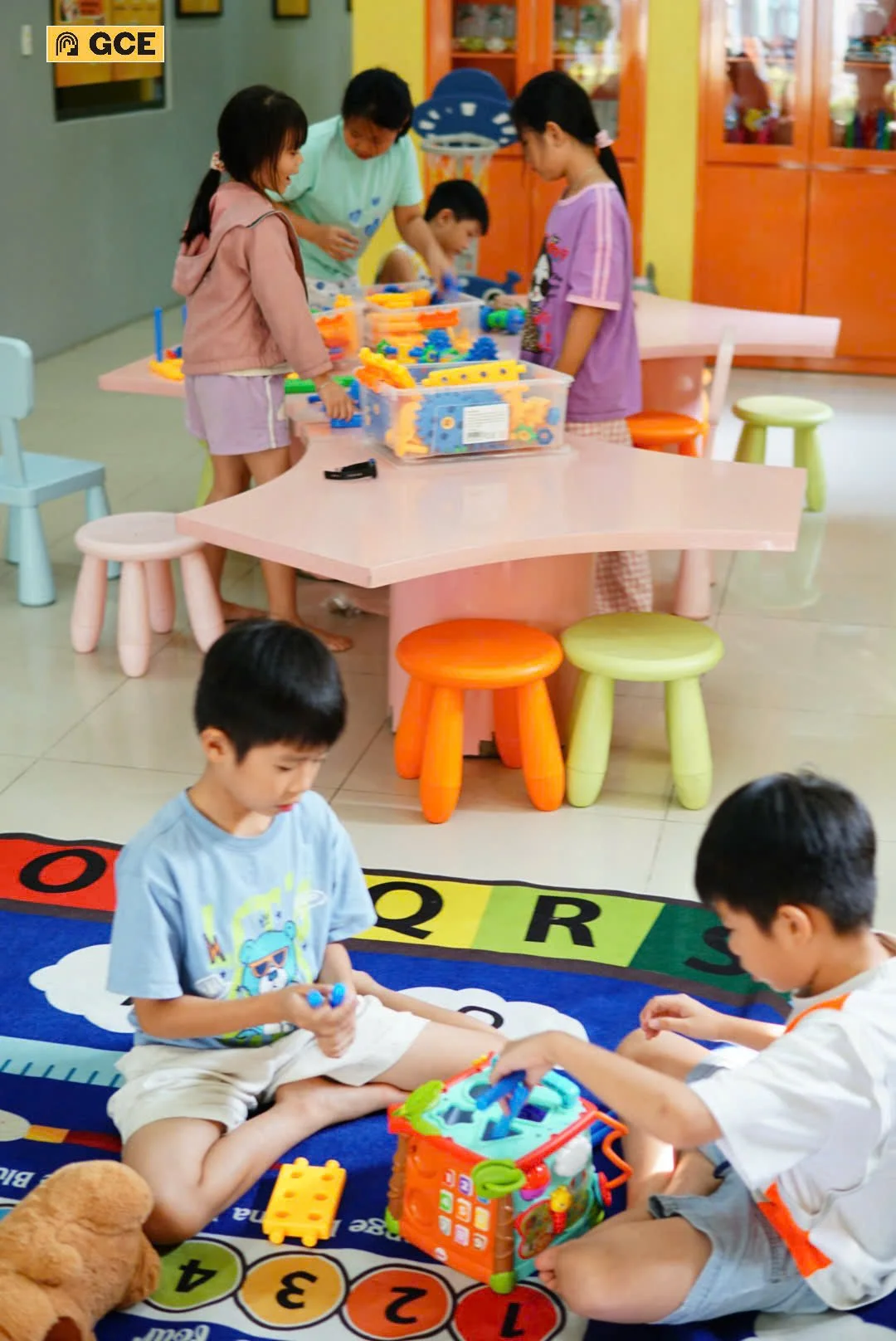 Children playing with building toys in a colorful indoor playroom, with a group of children at a pink table with plastic gears and building sets, and two children sitting on a multicolored learning carpet playing with a plastic stocking toys.
