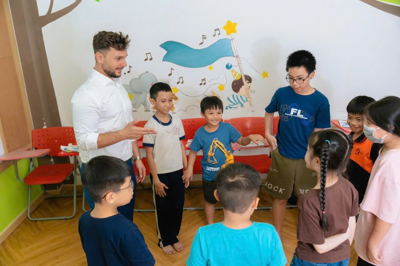 A man and children in a circle in a classroom with a wall mural of stars, flags, and kids playing, some children are smiling and engaging, one child is wearing a face mask.