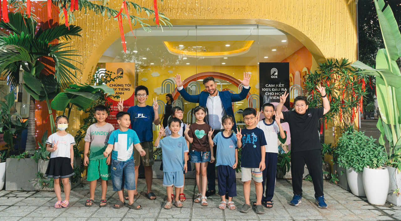 A group of children and two adults standing in front of a decorated building, some children making peace signs and raising their hands, with tropical plants on either side.