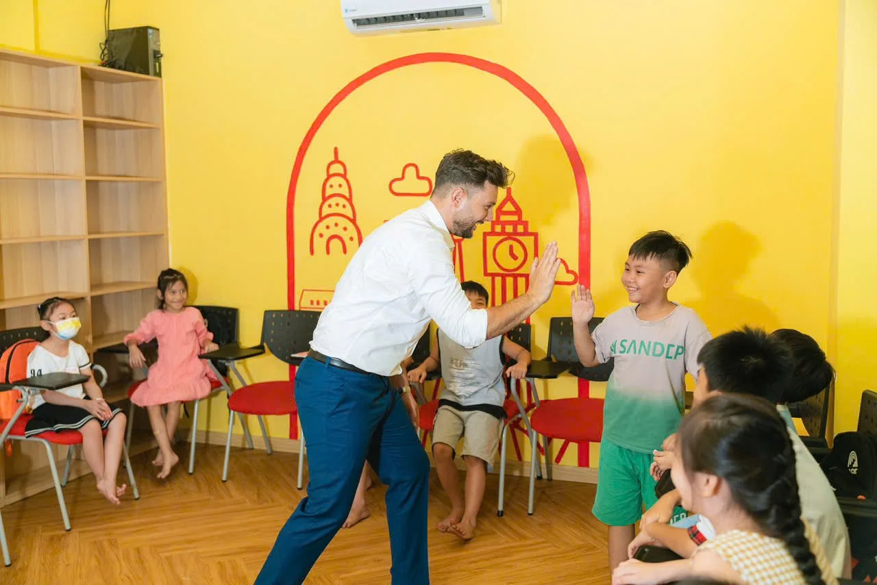 Young man high-fiving boy in a classroom with colorful yellow walls and children sitting in chairs.
