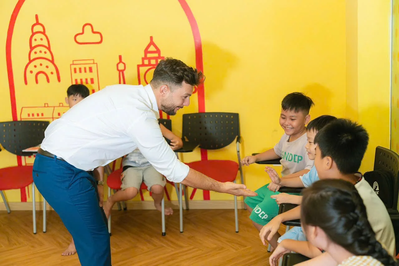 A man is interacting with a group of children seated in a row, all smiling and laughing in a colorful classroom with bright yellow walls and cityscape drawings.