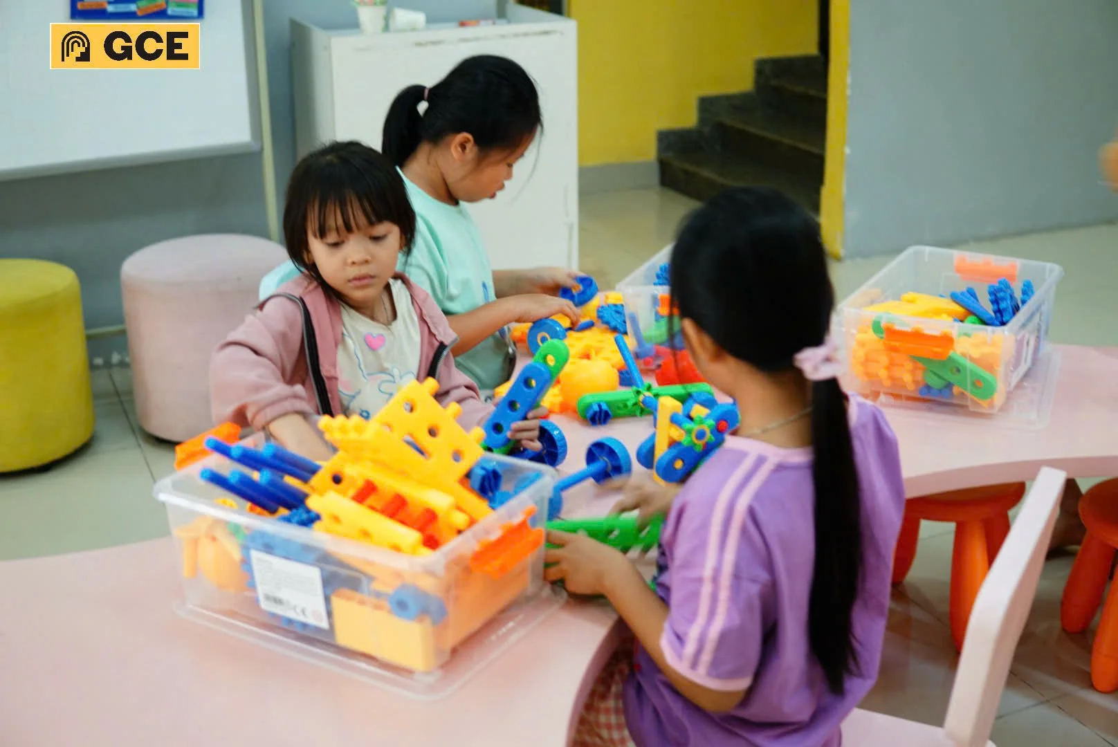 Three young girls are playing with colorful building blocks at a pink table in a room with yellow and gray walls. Two transparent plastic containers filled with various brightly colored plastic pieces are on the table.