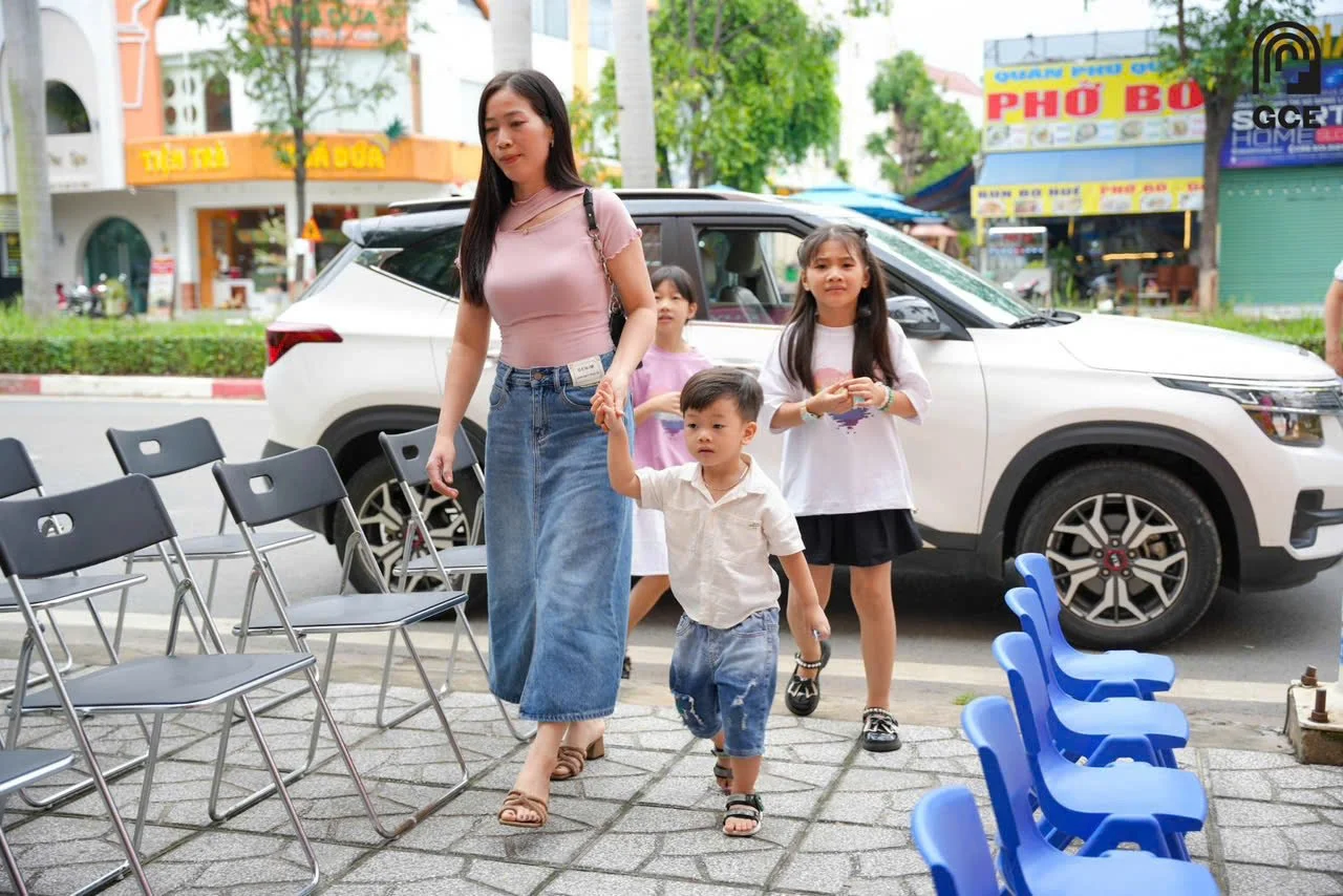 A woman and three children walking on a sidewalk in front of parked cars and colorful storefronts, with chairs lined up on the sidewalk.