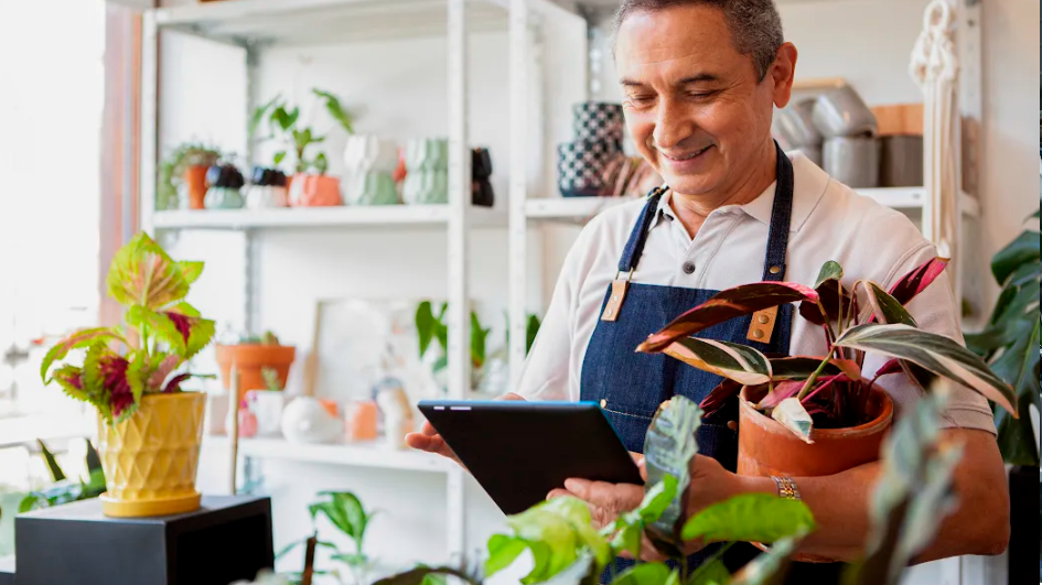 A man in a white shirt and apron smiling while looking at a tablet in a plant shop with various potted plants on shelves.