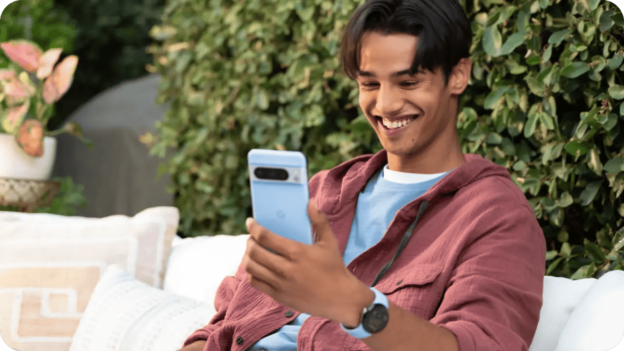 A young man sitting on an outdoor couch, smiling as he looks at his smartphone surrounded by greenery and potted plants.