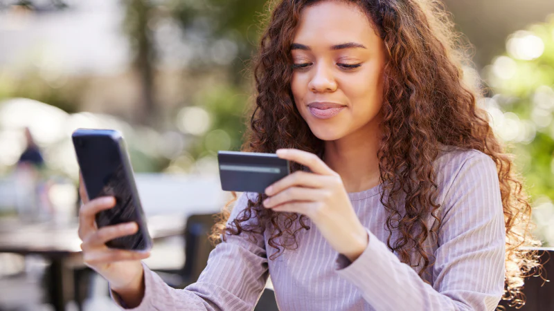 A young woman with curly hair is sitting at an outdoor cafe, looking at her smartphone and holding a credit card, possibly making an online purchase.