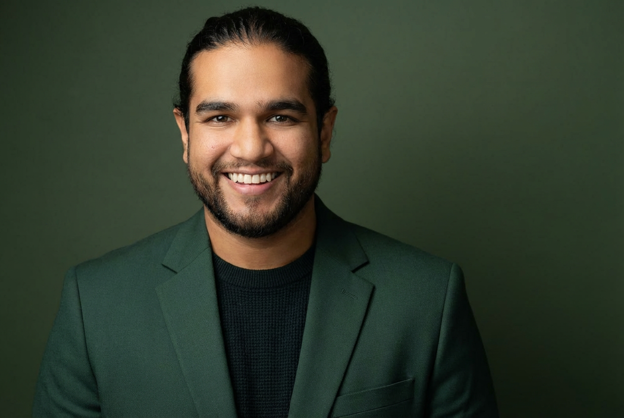 Headshot of a smiling man with dark hair and beard, wearing a dark green suit jacket and black shirt, against a green background.