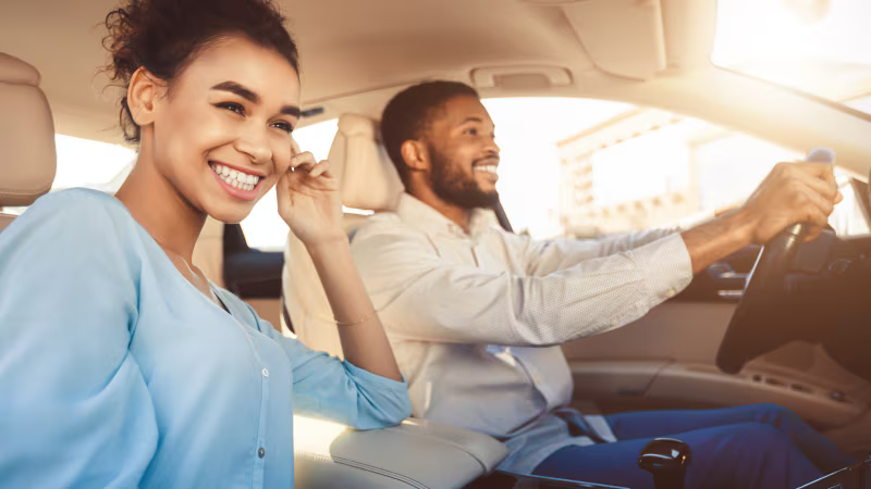 A woman in a blue blouse sitting in the passenger seat and smiling, while a man in a light shirt driving a car and smiling.