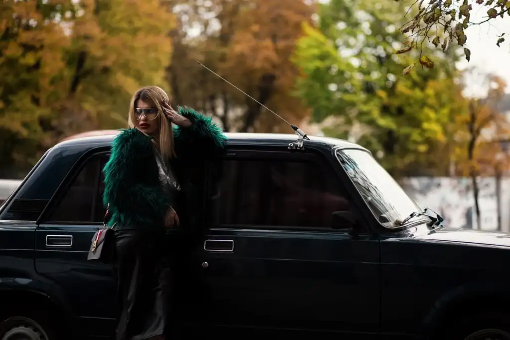 A woman in sunglasses and a dark green fur coat standing next to a black vintage car, with autumn trees in background.
