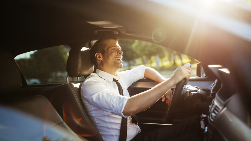 A man in a white shirt and black tie driving a car with a smile on his face during sunset.