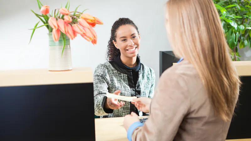 A woman with curly hair smiles while sitting at a reception desk, handing an ID card or document to another woman with long, straight, blond hair who is facing away from the camera. There is a vase of pink tulips on the desk and a potted plant in the background.