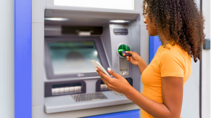 A woman using an ATM machine, holding a smartphone in one hand and pressing buttons with the other.