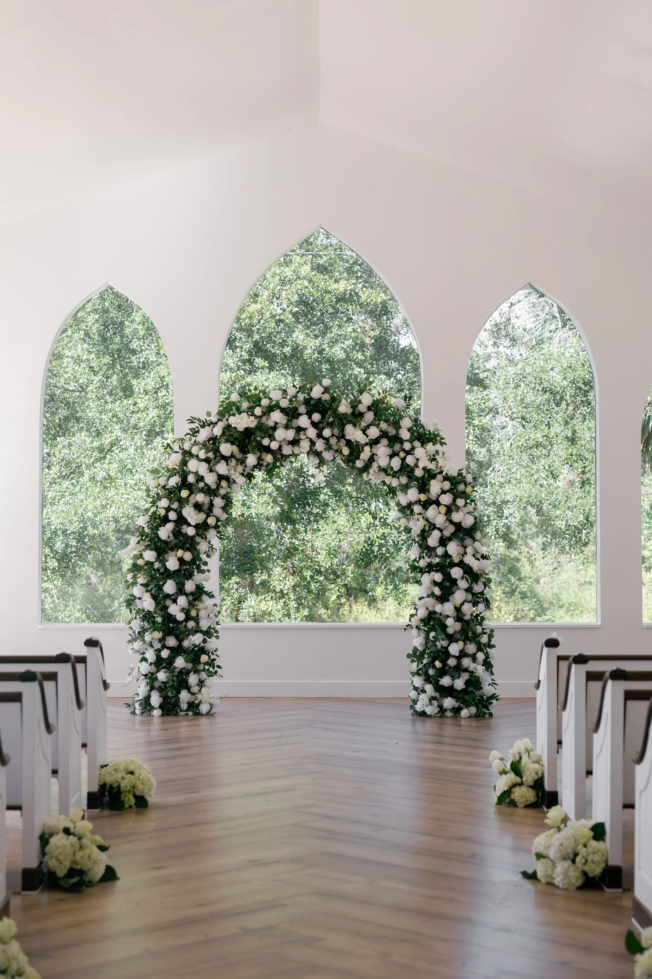 A wedding ceremony setup inside a white church with large arched windows, a floral arch decorated with white flowers and greenery, and white flower arrangements on the wooden floor along the aisle.