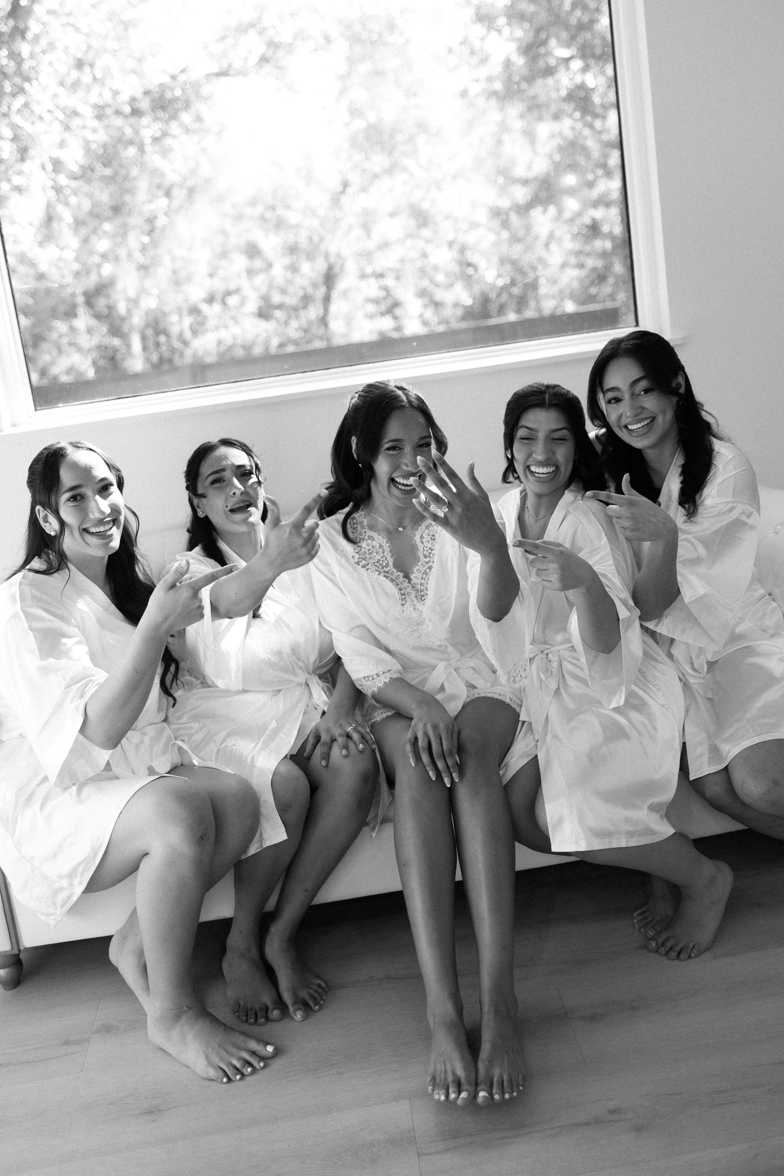 A group of six women in white robes sitting on a couch, laughing and pointing at a woman showing her wedding ring, in a room with a large window and natural light.