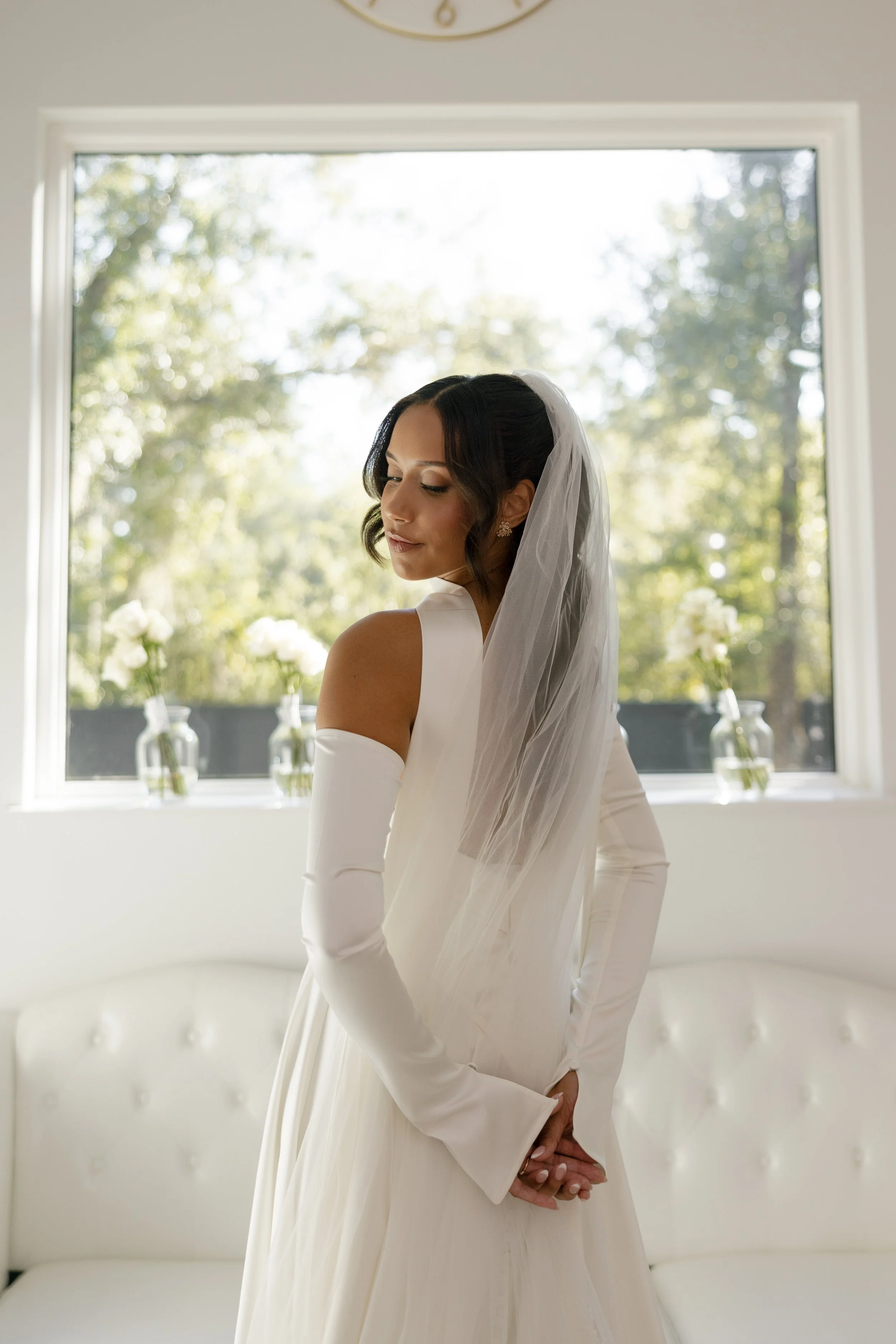 A bride wearing an off-shoulder white wedding gown and long gloves, with a veil, standing in a bright room with a large window and vases of white flowers in the background.