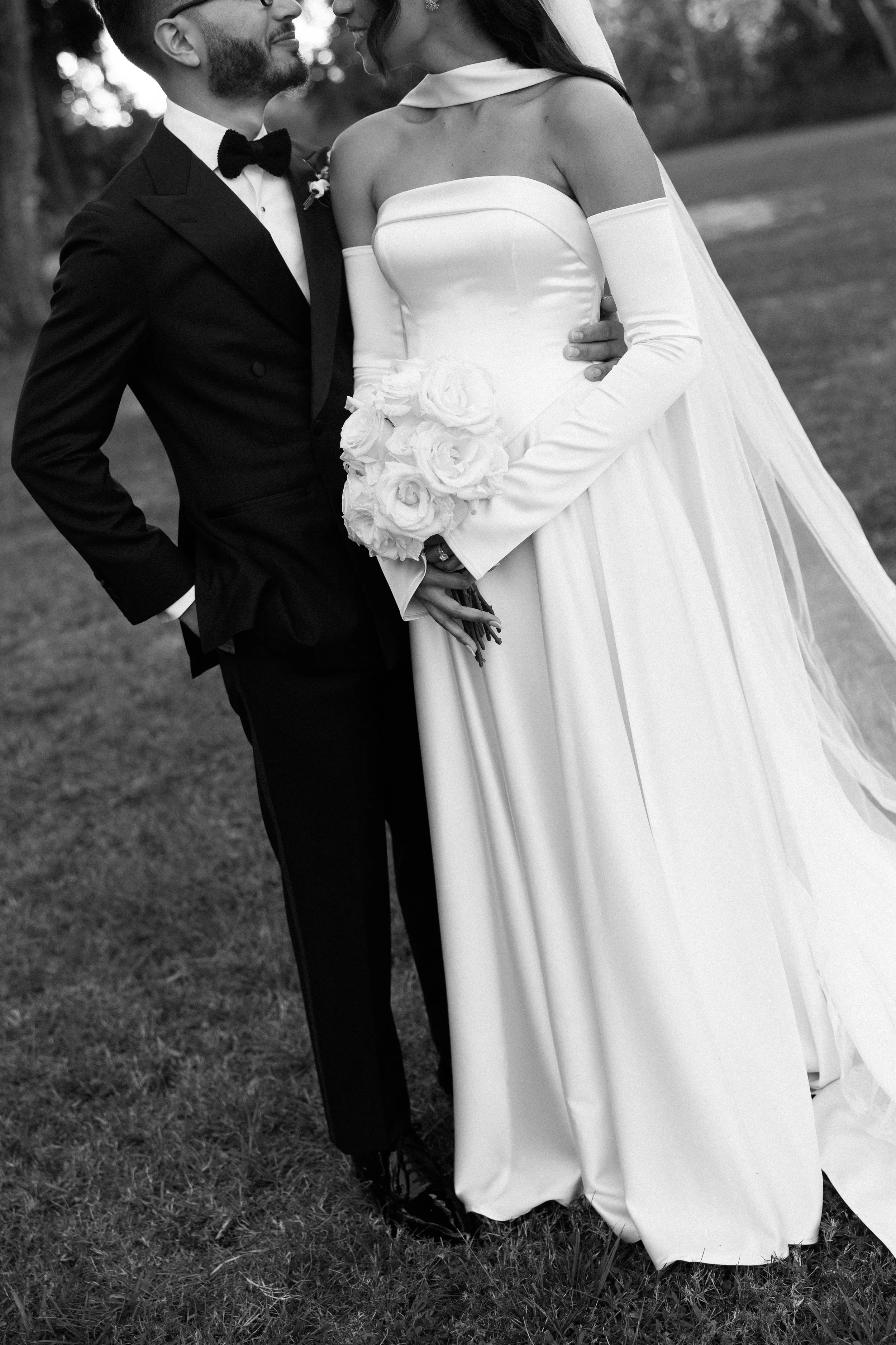 Black and white photo of a bride and groom outside on grass, close together, with the bride holding a bouquet of roses.