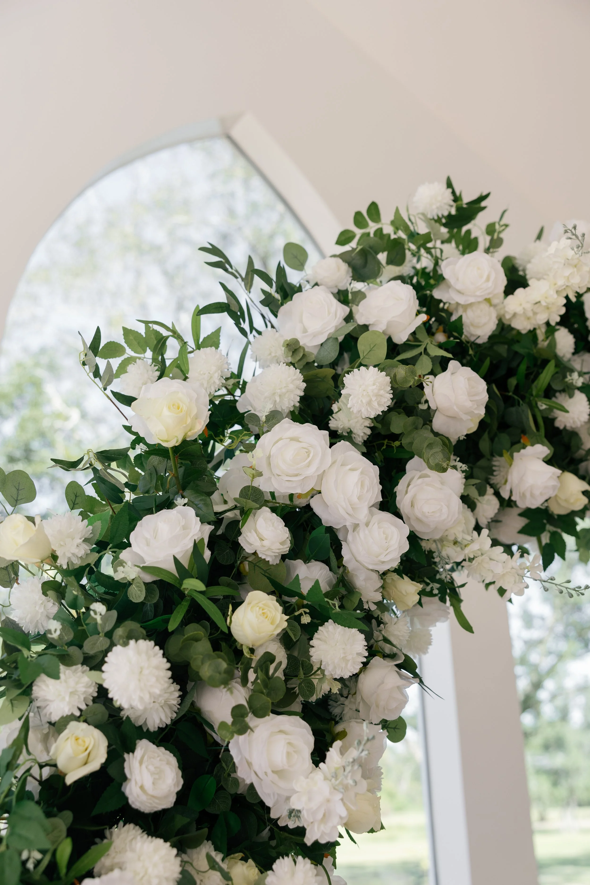 White floral arrangement with roses, dahlias, and greenery near a window in a bright room.
