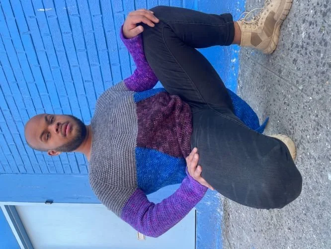 Alexander Reynoso of AlexCreates seated outdoors in a gray, blue and purple color-block sweater