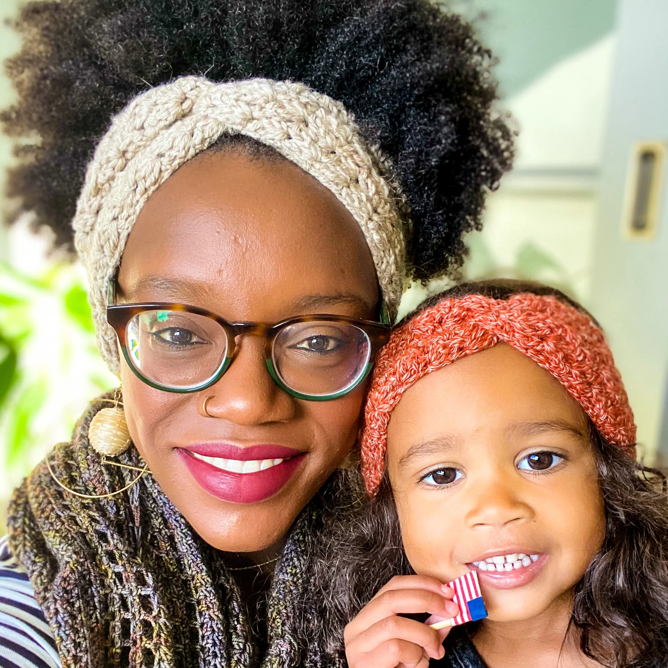 Woman and young child smiling in matching Cara Earwarmers, hers in oatmeal and the child’s in coral, each with a twist at the front. Free crochet earwarmer pattern.