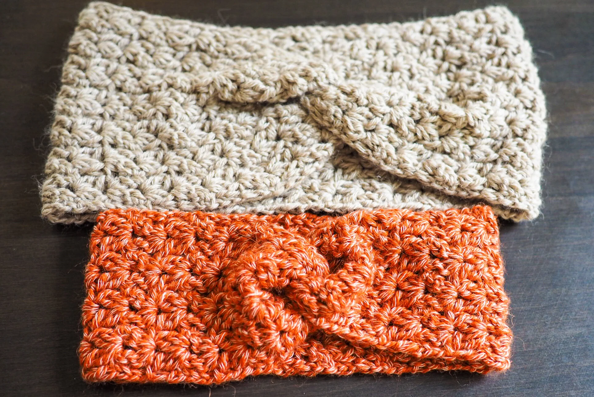 Two Cara Earwarmers laid flat, an adult size in oatmeal and a child size in coral, showing the twist and textured stitches. Free crochet earwarmer pattern.