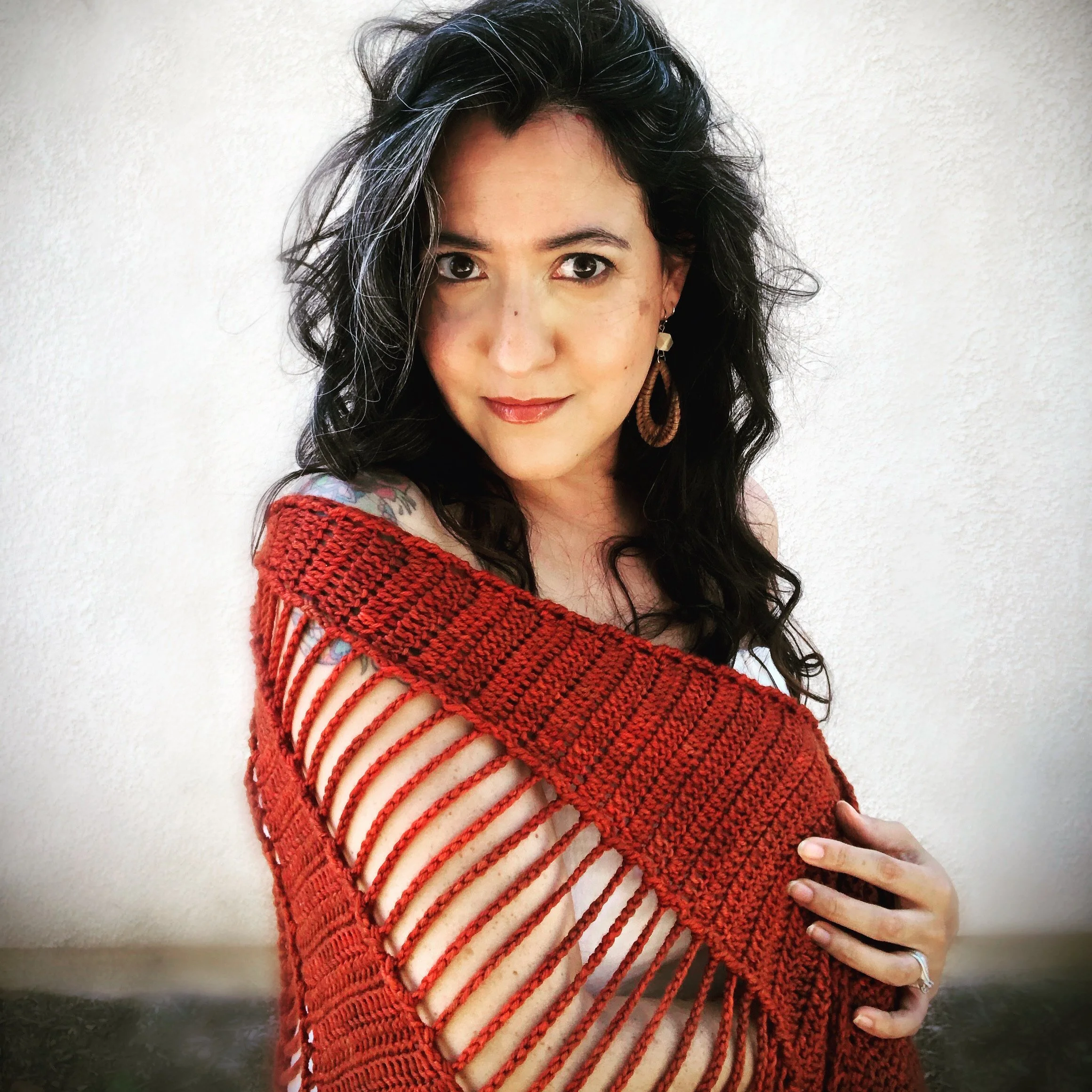 Woman wearing a red, knitted, open-stitch sweater, posing against a white background, with long dark hair and hoop earrings.