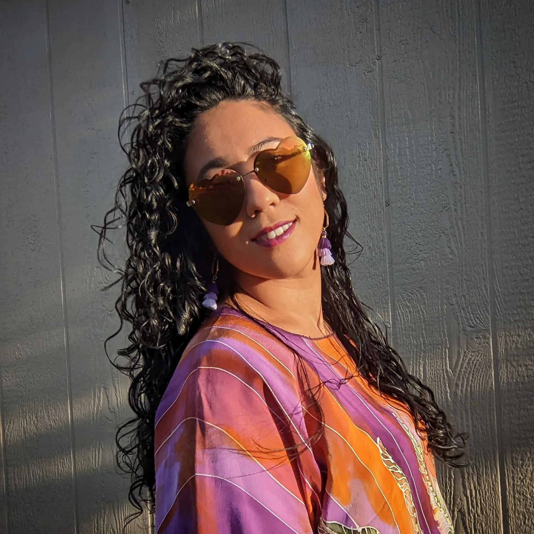 Woman with curly hair wearing sunglasses and colorful blouse, standing against a wooden wall.