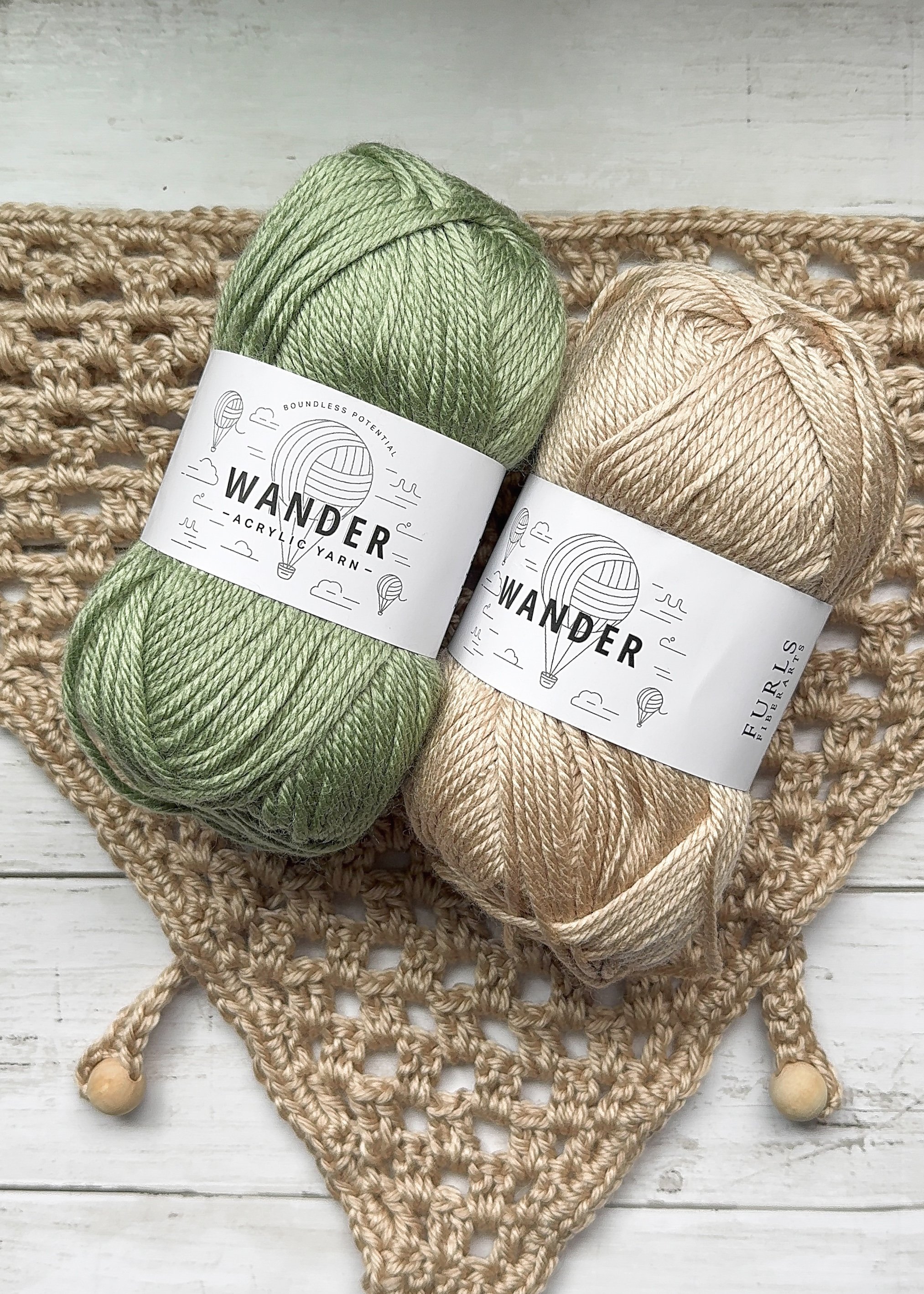 Sage green and tan skeins of Furls Wander yarn resting on a tan crochet bandana with wooden bead ties. Easy Drape Bandana, a free crochet pattern.