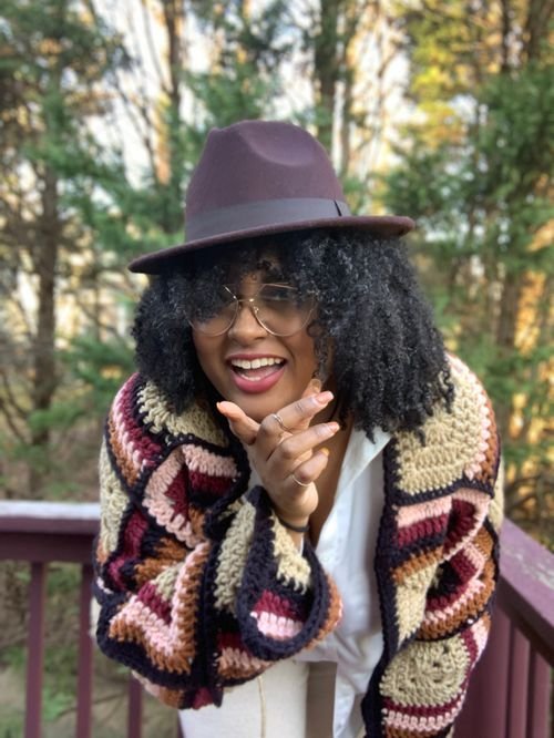 India Yasmein of Stitch'd by India in a burgundy hat and a granny square crochet cardigan