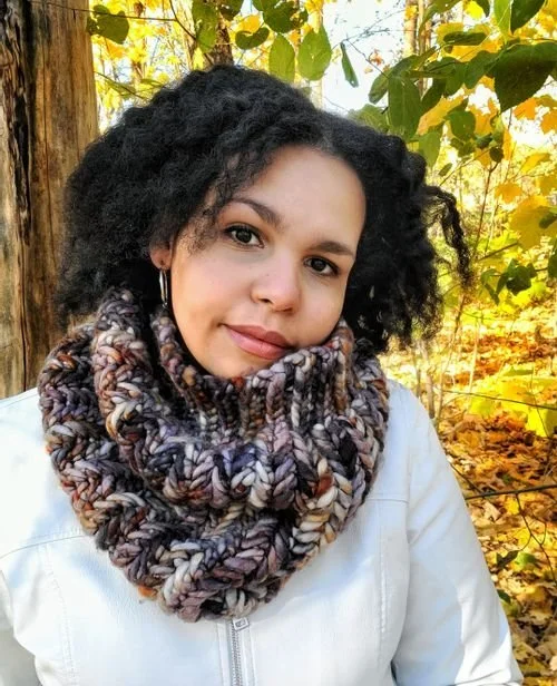Roxanne W of eat.wander.crochet in a chunky brown and cream cowl among fall leaves