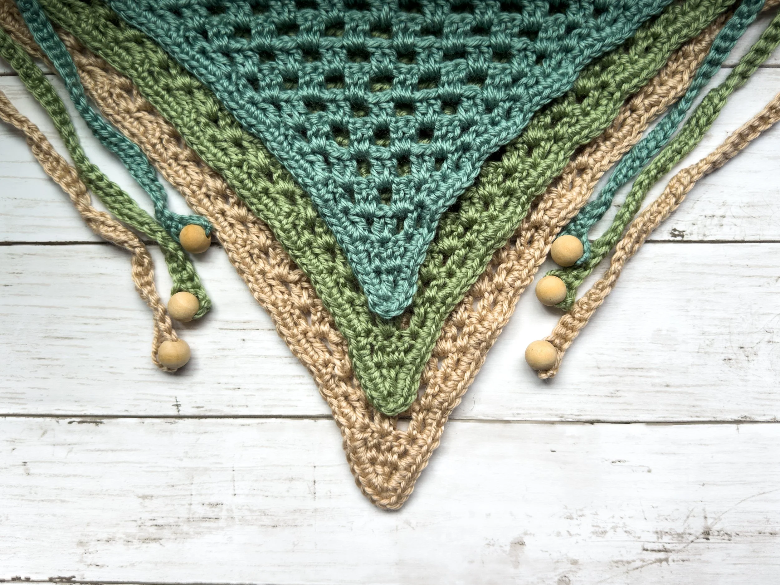 Three Easy Drape Bandanas in teal, sage and tan, with wooden bead ties, laid flat on wood. Free beginner crochet bandana pattern.