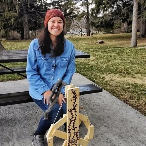 Raeli Masina of Erieli Knits in a rust beanie and denim shirt, seated beside a wooden yarn swift