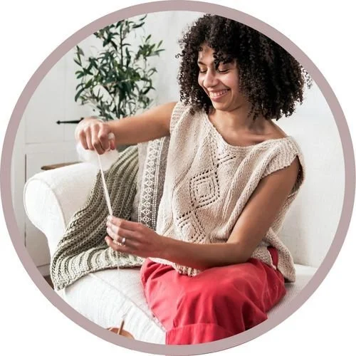 Lauren McElroy of Mother of Purl knitting on a sofa in a cream lace-panel knit top