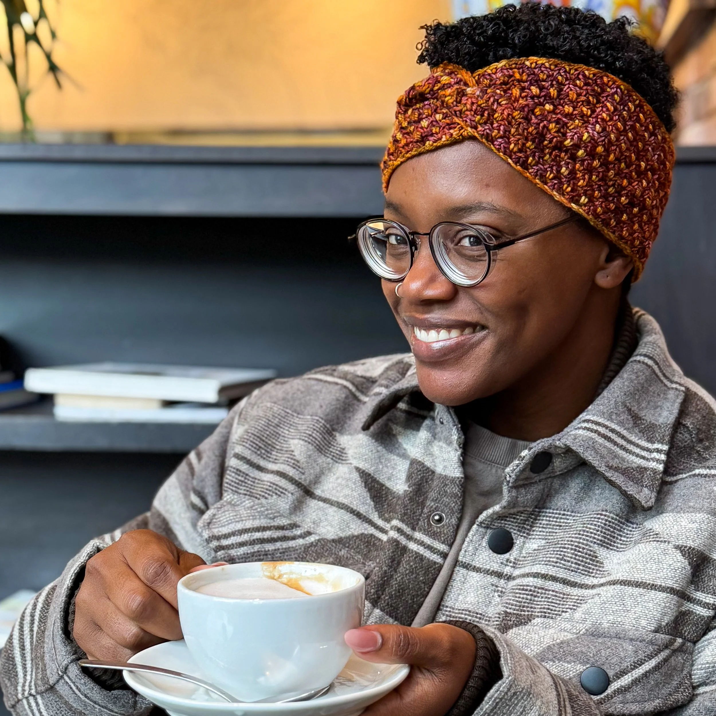 Woman smiling over a cup of coffee, wearing a rust and gold textured crochet headband with a twist. Trifle Headband, a free beginner crochet pattern.