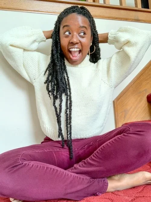 Alexandrea Owens of Craftedbyalliejean sitting cross-legged in a cream knit sweater, grinning