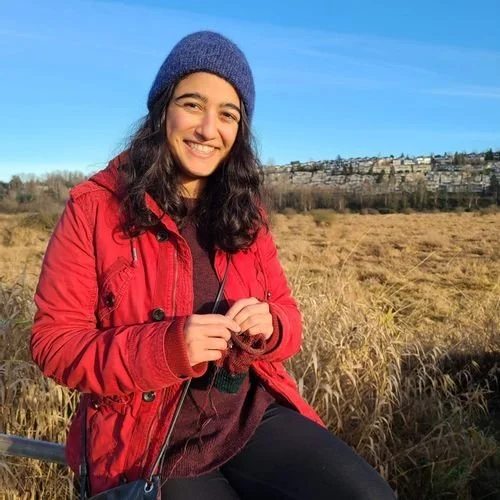 Natasha Pekurel of Northern Knits and Purl knitting outdoors in a navy beanie and red jacket