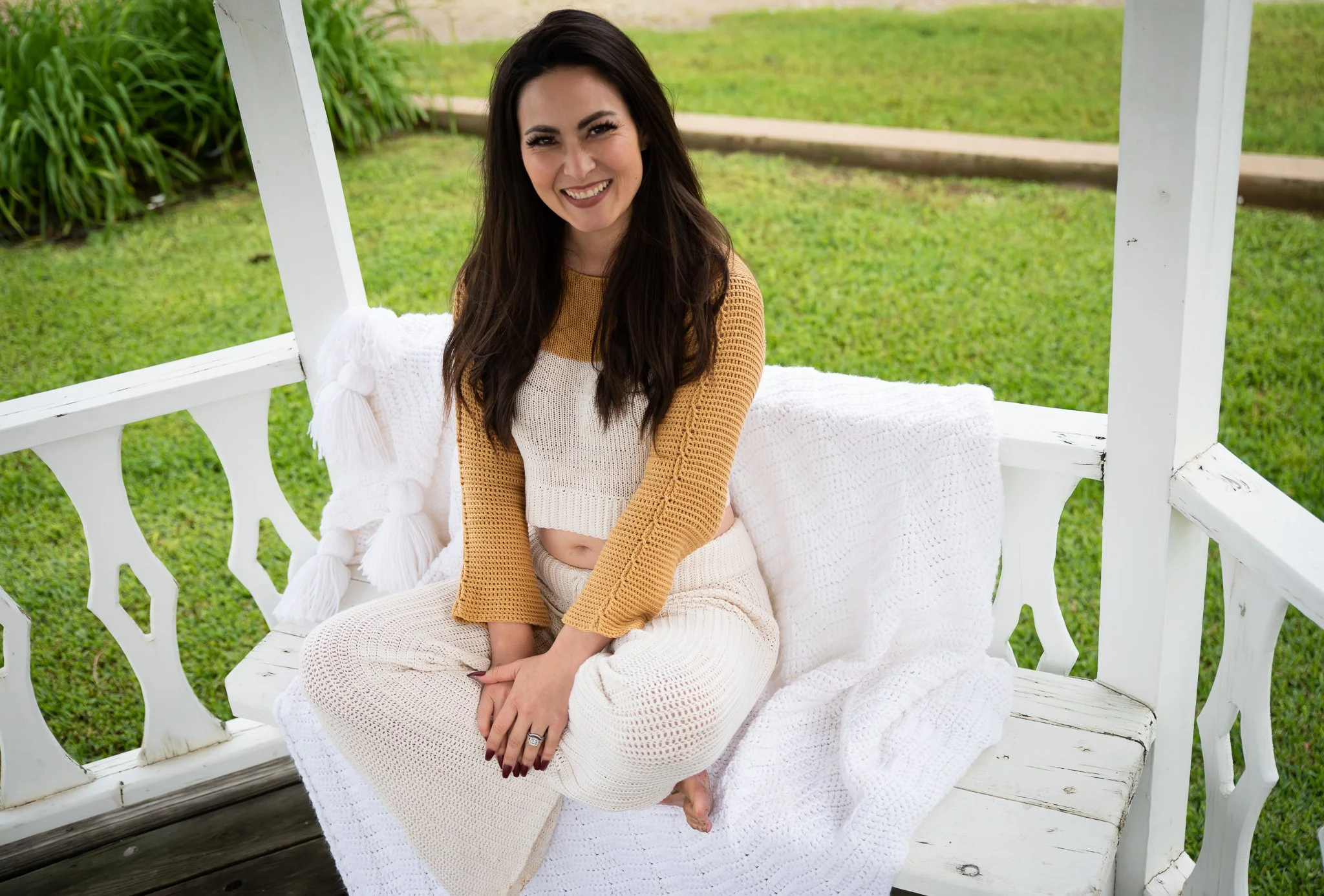 An asian woman sitting on a white wooden bench, wearing a yellow and white crochet outfit, smiling, with a green grass background.