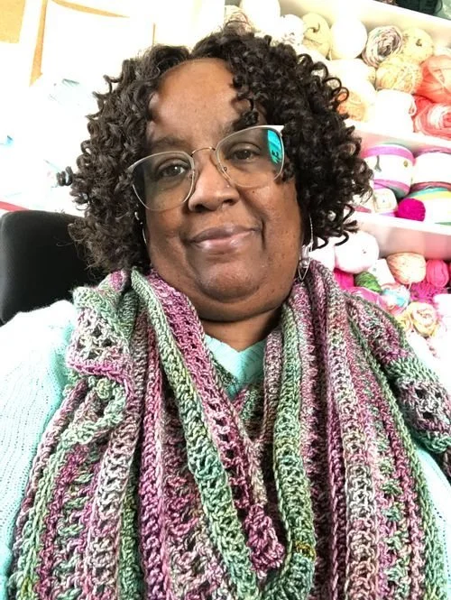 LaVonne Switzer of LS Crochet Designs in a pink, green and cream crochet scarf, with yarn shelves behind
