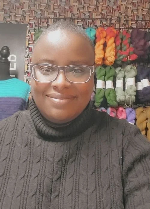 Lakisher Hurst of Sassy Black Yarns smiling in a cable knit turtleneck, with yarn shelves behind
