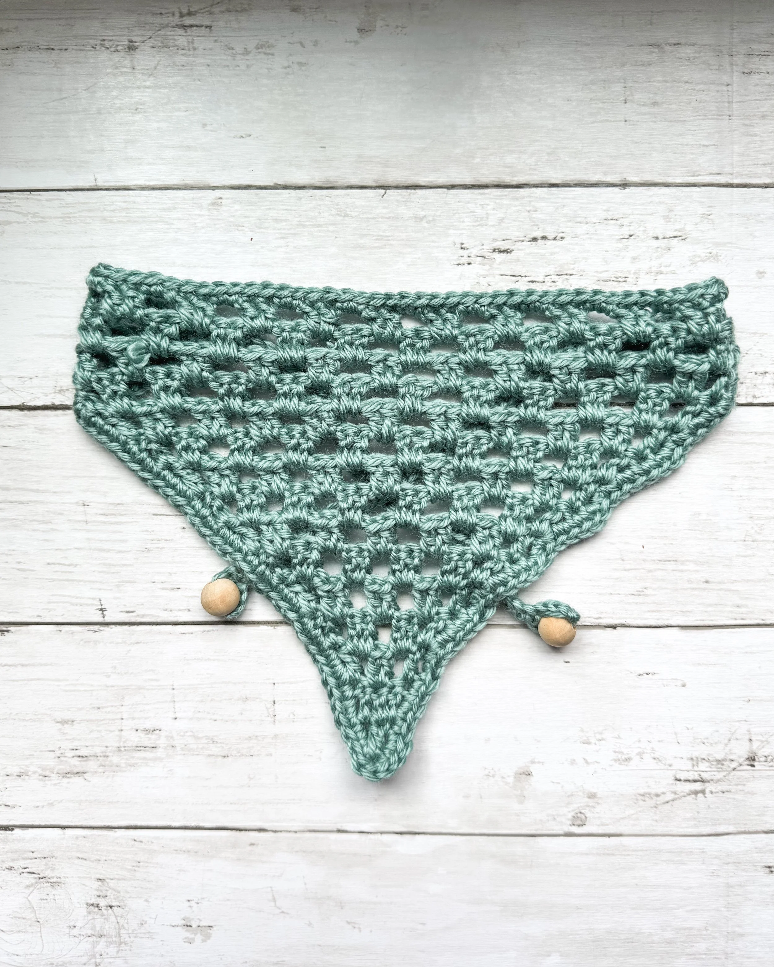 Teal Easy Drape Bandana laid flat on white wood, showing the open mesh triangle and wooden bead ties. Free beginner crochet bandana pattern.