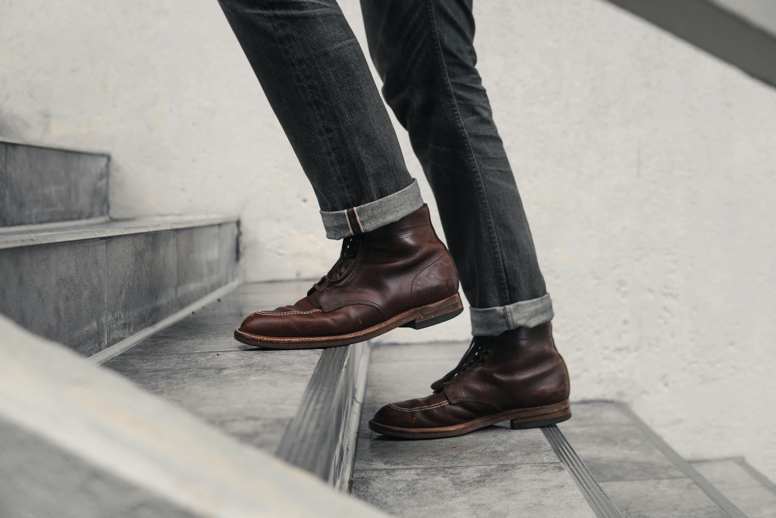 Close-up of a person's legs wearing dark jeans with cuffs rolled up, brown leather boots, and stepping on a staircase with grey tiles and metal trim.
