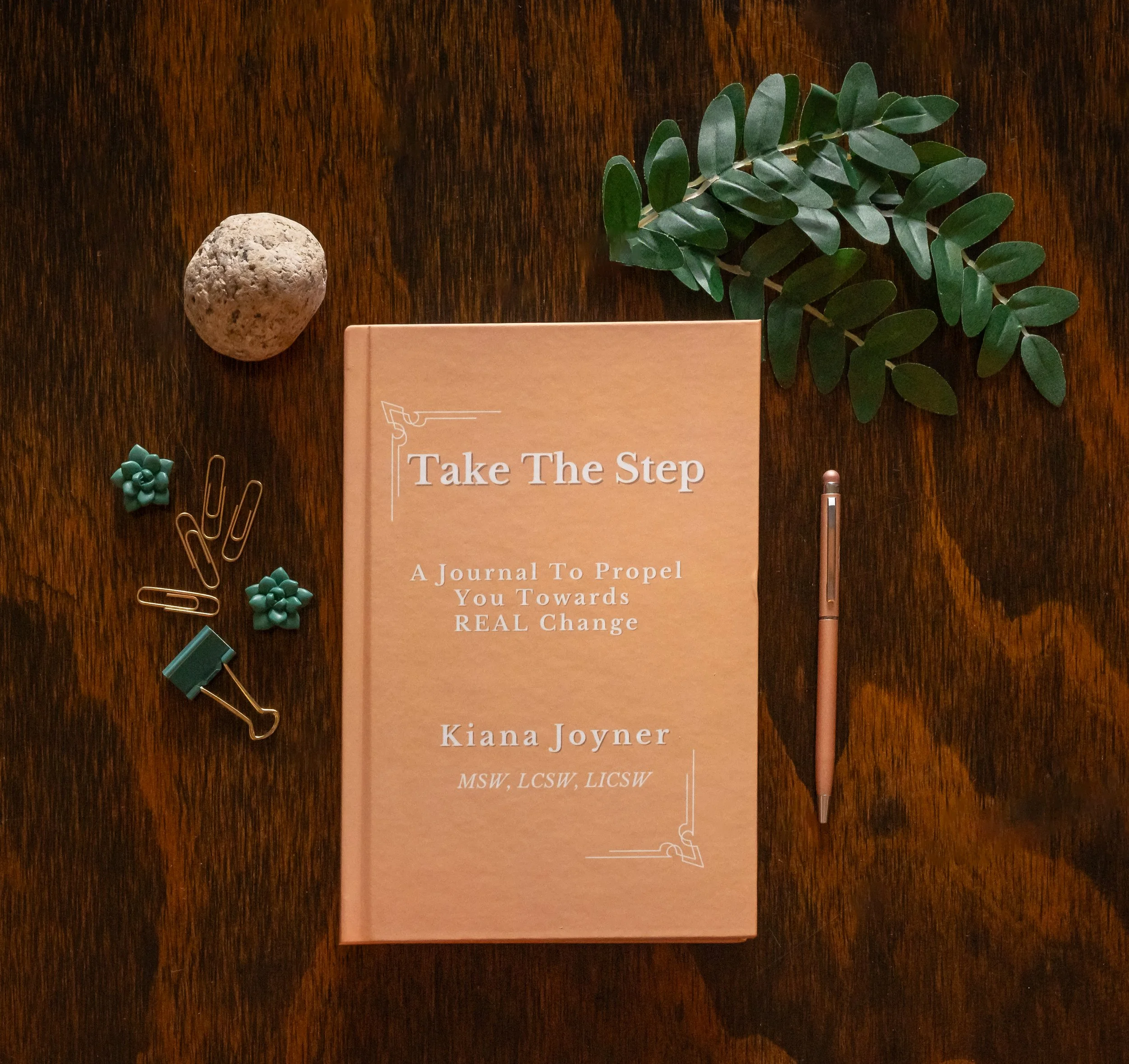 A peach-colored journal titled 'Take The Step' with the subtitle 'A Journal To Propel You Towards REAL Change' by Kiana Joyner, surrounded by a small green plant, a gray stone, blue decorative clips, a gold paperclip, and a rose gold pen, all on a wooden surface.