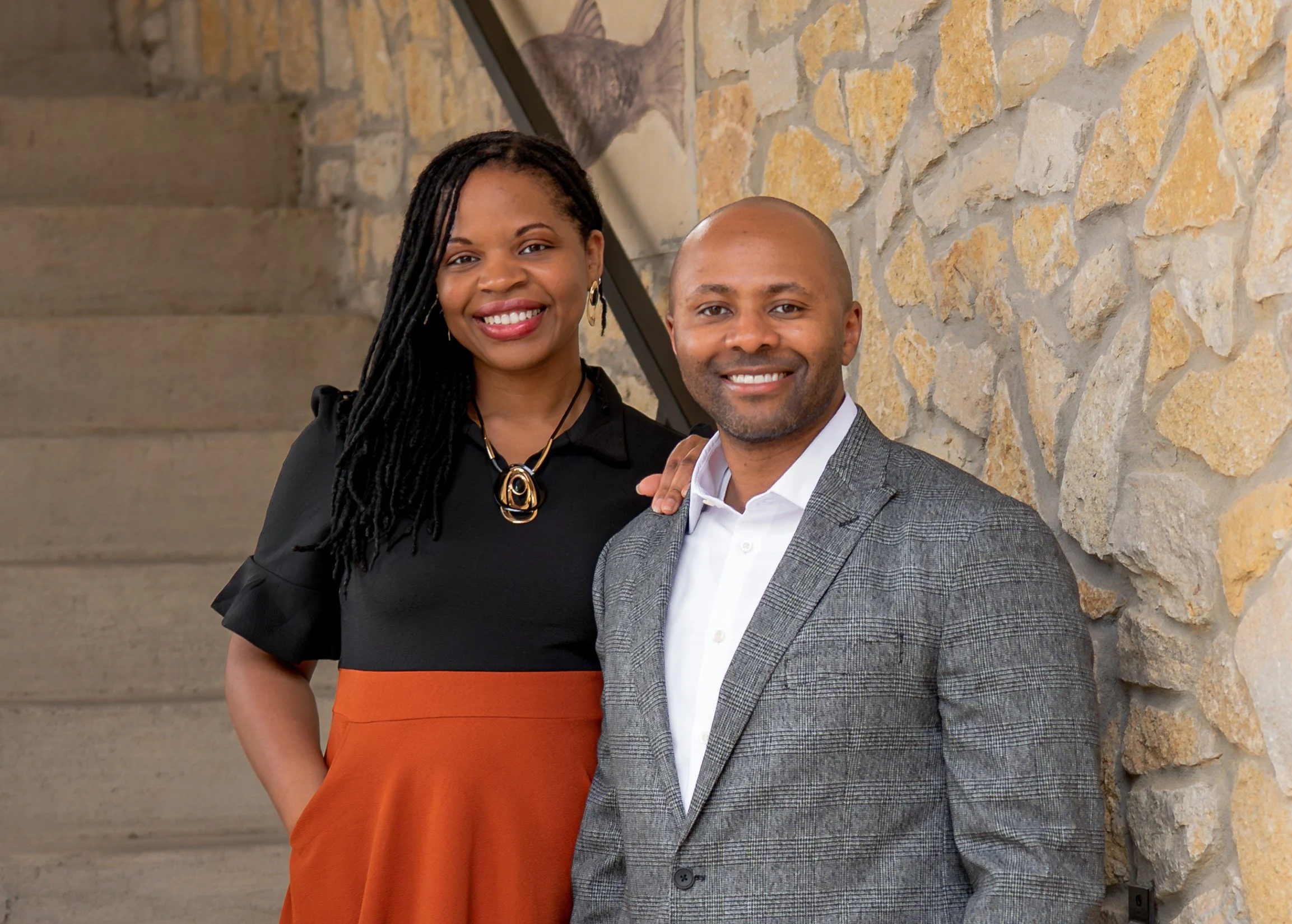 A woman and a man standing close together indoors, smiling, with a stone wall background. The woman has long dreadlocks and is wearing a black dress with orange accents and a large necklace. The man is bald, wearing a gray checked blazer over a white shirt.