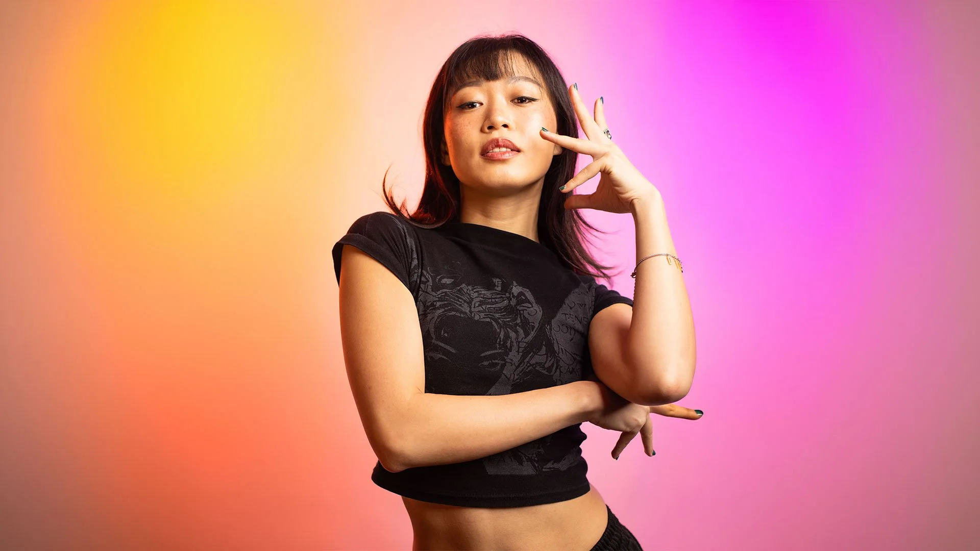 A young woman with dark hair wearing a black t-shirt with a graphic design, standing with her arms crossed and one hand near her face, in front of a colorful gradient background blending orange, yellow, pink, and purple.