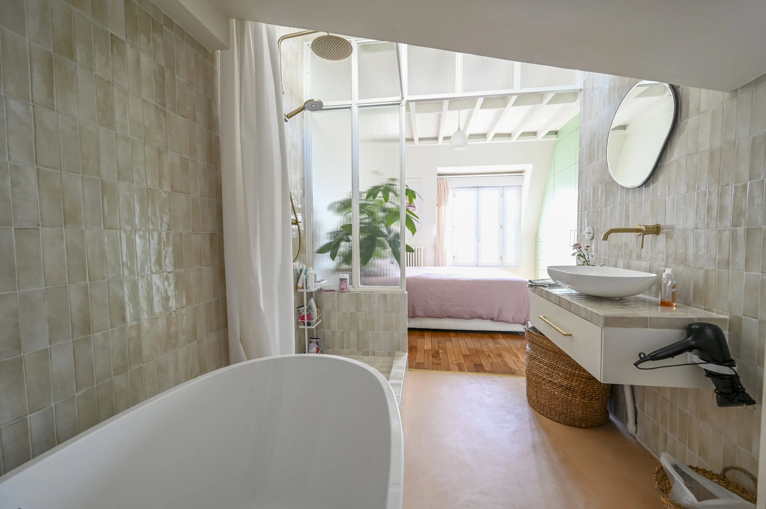 A bright bathroom with beige tile walls, a white bathtub, a shower area separated by a glass partition, and a vanity with a vessel sink. There is a mirror above the sink, a wicker basket, and a large window bringing in natural light. A bed with pink 