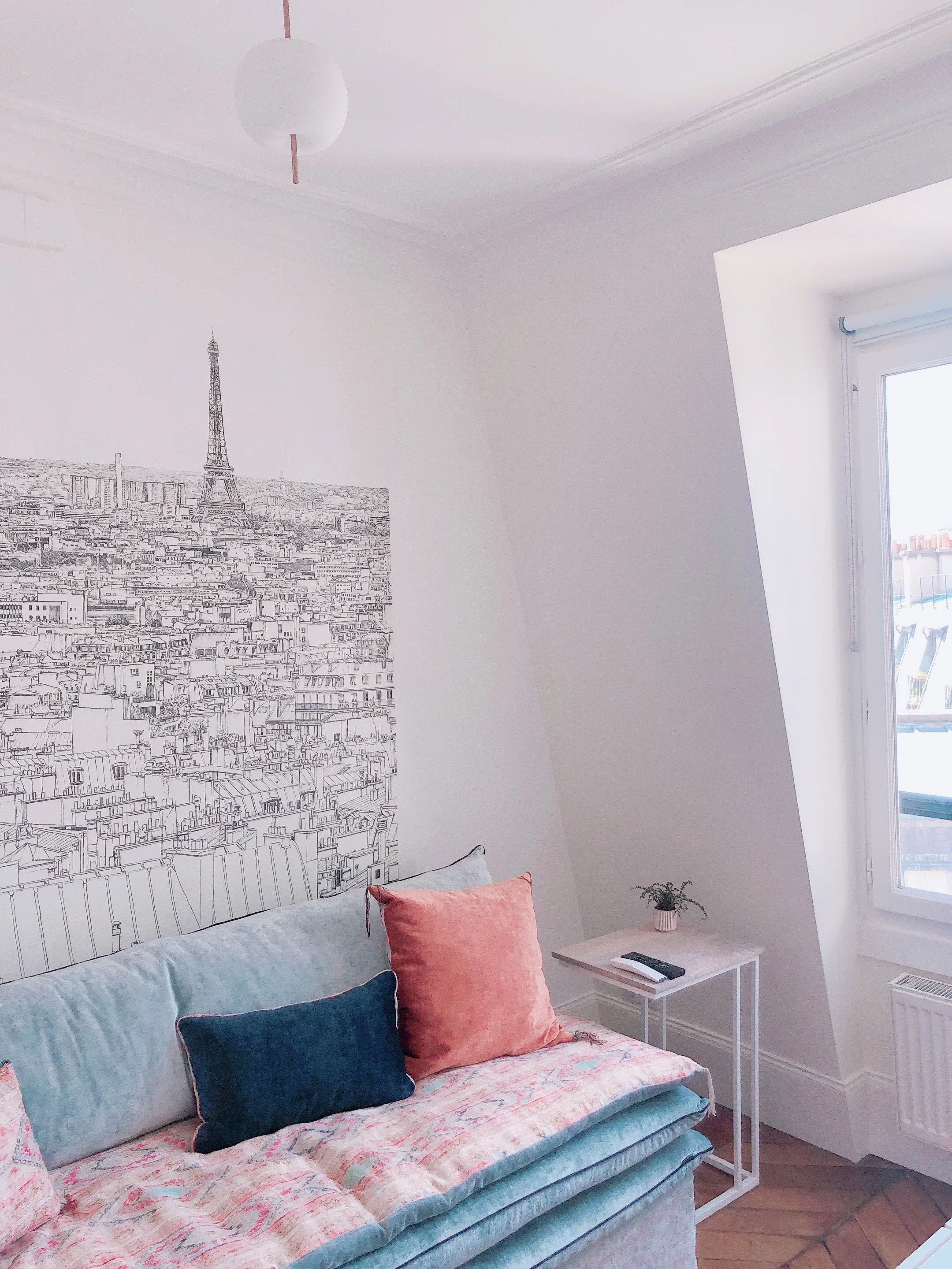 A cozy living room corner with a light-colored sofa, featuring three cushions in peach, navy, and coral. Next to the sofa is a small white side table with a remote and a cell phone, and a potted plant rests on it. The wall behind the sofa displays a 